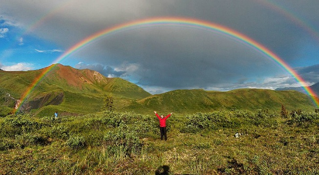 person standing on grassy mountain under double rainbow